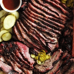 Smoked beef brisket sliced and garnished with pickles, a small bowl of BBQ sauce, and sliced peppers.